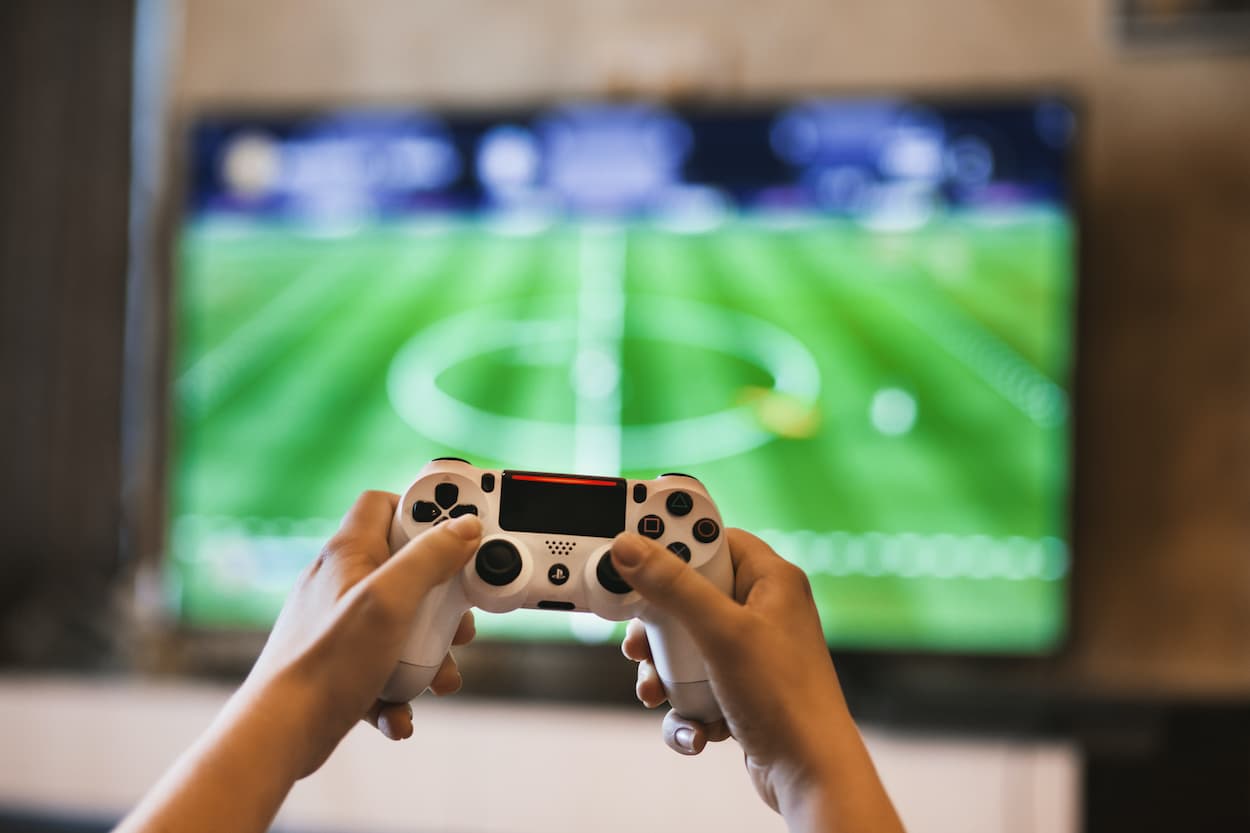 Pessoa jogando no console com tv ao fundo em partida de futebol