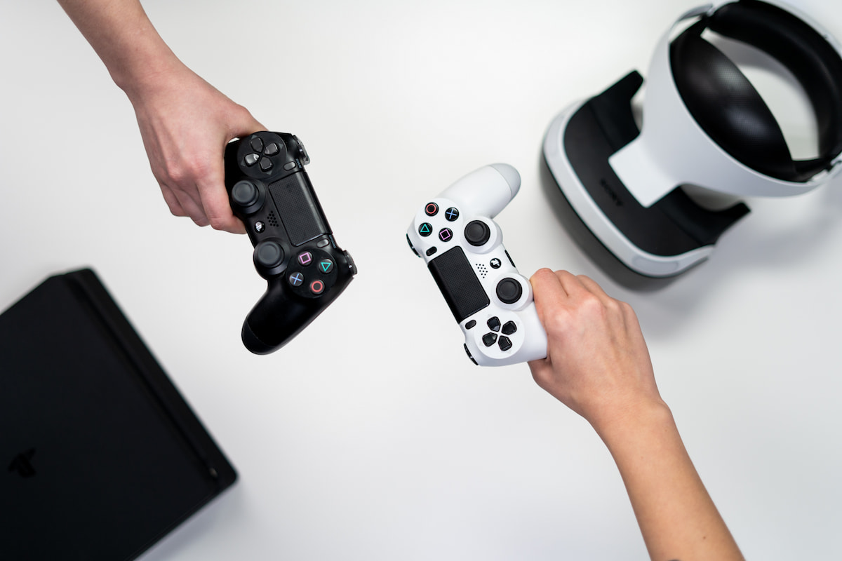 Duas pessoas segurando controles do PS4 pretos e brancos, com acessórios do console ao fundo desfocados