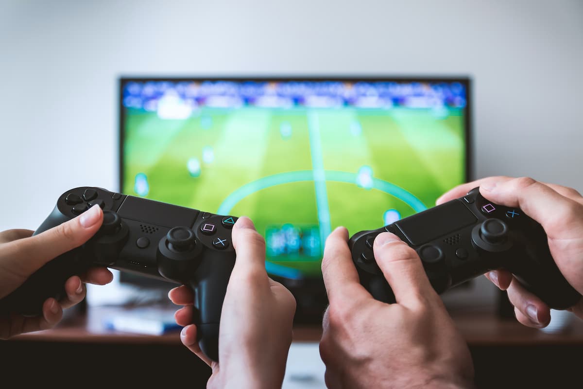 Duas mãos segurando controle de PS4 jogando um jogo de futebol no fundo