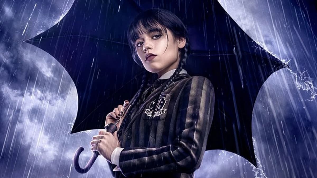 Personagem Wandinha da Netflix, interpretada pela atriz Jenna Ortega, menina com maquiagem escura, tranças, roupas de colegial preta com guarda-chuva aberto lhe protegendo da chuva.