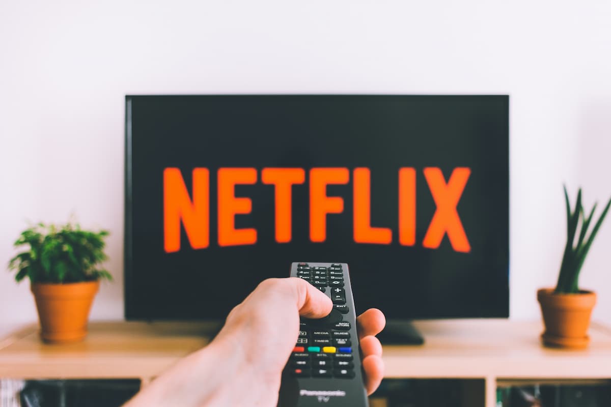 Mão com controle apontando para TV com logo da Netflix na tela.