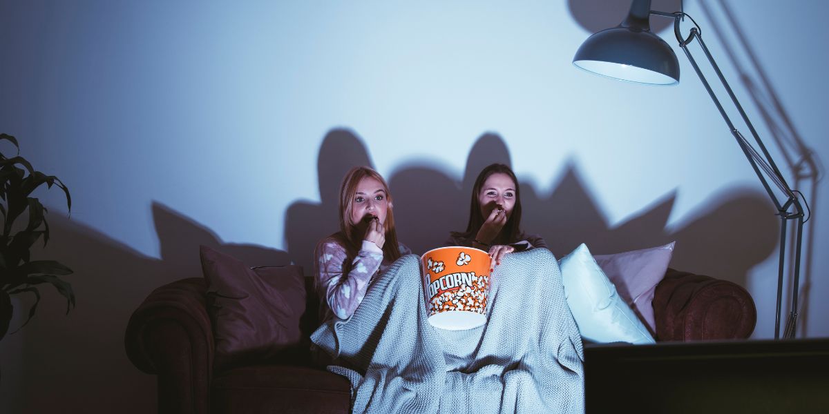 Duas mulheres sentadas em um sofá, enroladas com lençol e comendo pipoca enquanto assistem a um filme de terror.