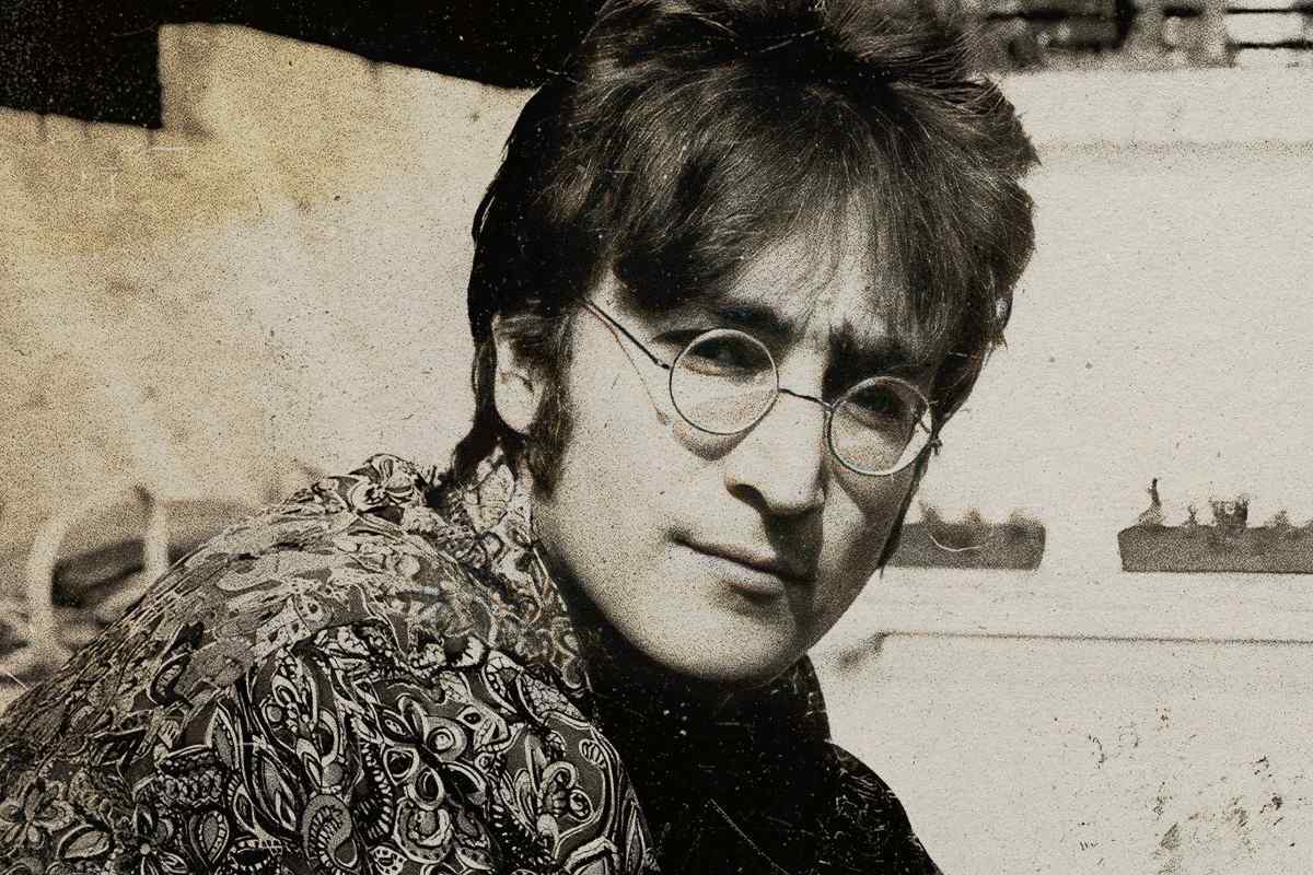 John Lennon. Foto antiga do cantor John Lennon. Nela, ele está com seus clássicos óculos redondos e uma camisa de estampa.