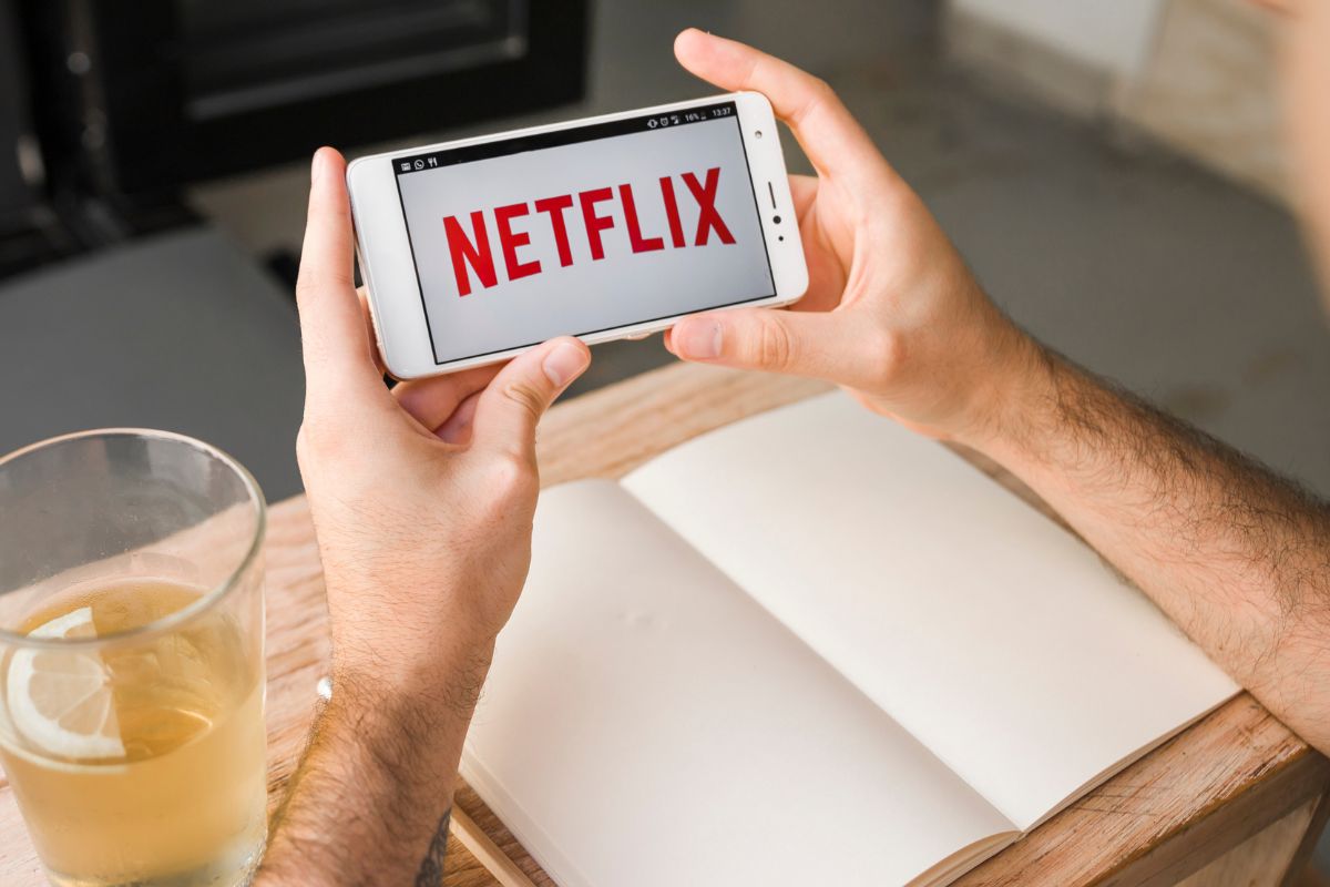 Homem com os braços apoiados em uma mesa de madeira, próximo a um copo de água com uma rodela de limão e de um caderno em branco, segurando um celular com o nome da Netflix na tela.