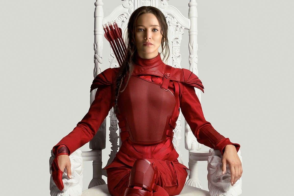 Jogos Vorazes. Pôster de divulgação do filme Jogos Vorazes. Nele, a protagonista está vestida vermelho, sentada em um trono todo branco e com um semblante sério.