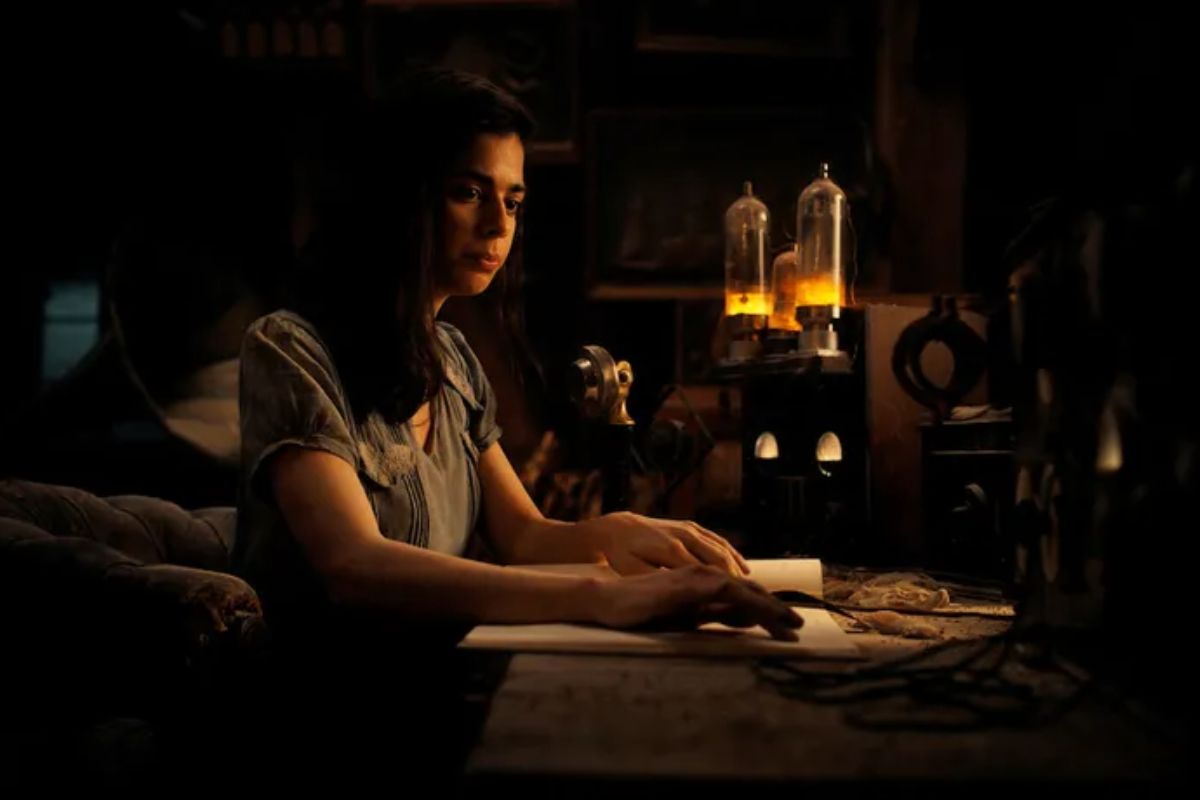 Toda Luz que Não Podemos Ver. Cena do filme Toda Luz que Não Podemos Ver, em que a protagonista está sentada em uma mesa, escrevendo em um papel em um ambiente escuro, à luz de algumas velas.