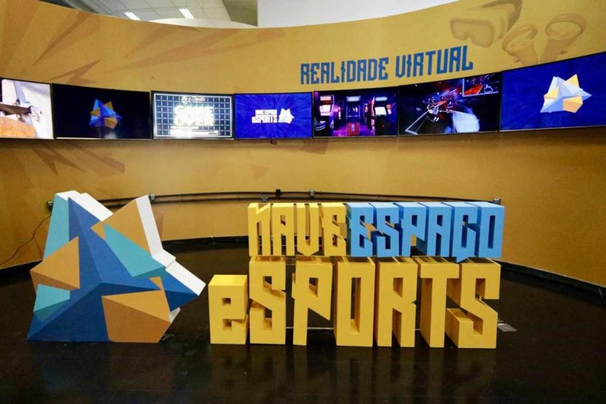 Foto da primeira Arena Gamer Pública do Brasil. Ao fundo, televisões estão expostas numa parede pintada de amarelo. Em destaque está um escrito Nave Espaço eSports.